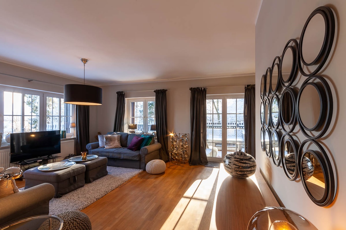 A spacious living area features large windows allowing soft natural light. Comfortable sofas are arranged to promote relaxation, complemented by decorative mirrors on the wall. A flat-screen television is positioned nearby, with a cozy rug placed on the wooden floor, enhancing the inviting atmosphere.