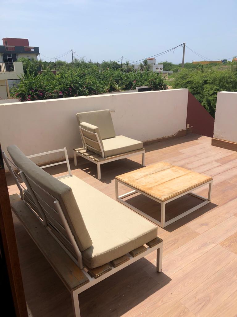 A private outdoor seating area features two light-colored lounge chairs and a wooden coffee table. The space is surrounded by a white wall and offers a view of greenery, enhancing the open, relaxed feel of the setting.