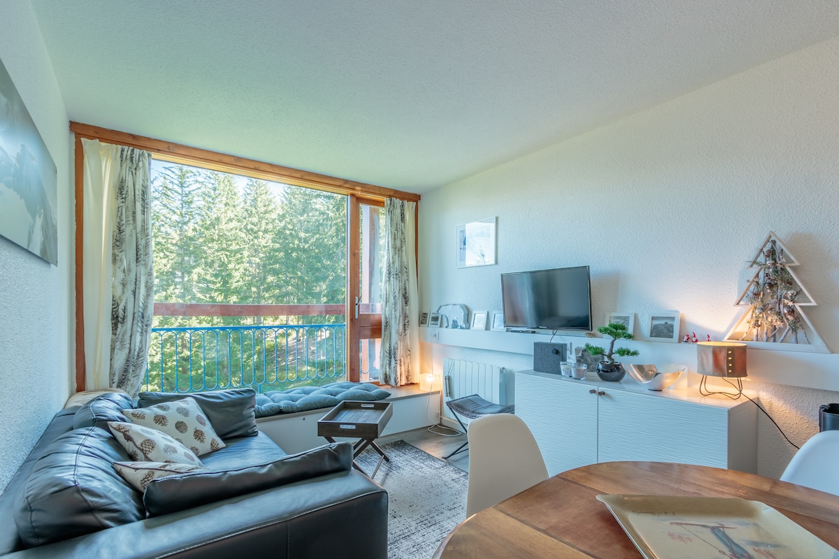 A comfortable living space features a dark leather sofa and a circular wooden dining table. A large window allows natural light to illuminate the room, revealing views of the surrounding trees. A TV is positioned on a white media unit with decorative elements placed nearby.