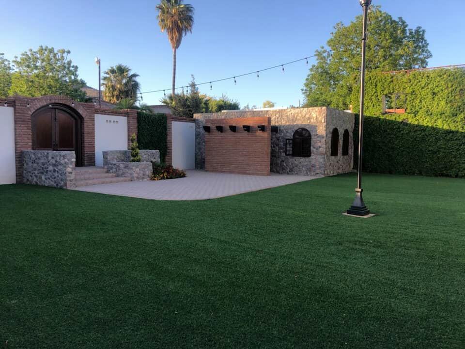 A spacious outdoor area is presented, featuring a well-maintained lawn bordered by lush greenery. Stone structures and a brick building create an inviting atmosphere. A decorative lamp post adds to the charm while string lights and vibrant flowers enhance the outdoor setting.