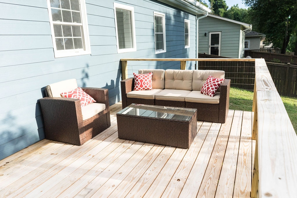 A private deck features a woven outdoor seating arrangement with a spacious sofa and lounge chair, adorned with decorative red pillows. A glass-top coffee table complements the setting. The deck has a wooden surface and is bordered by a natural landscape, providing a sense of seclusion.