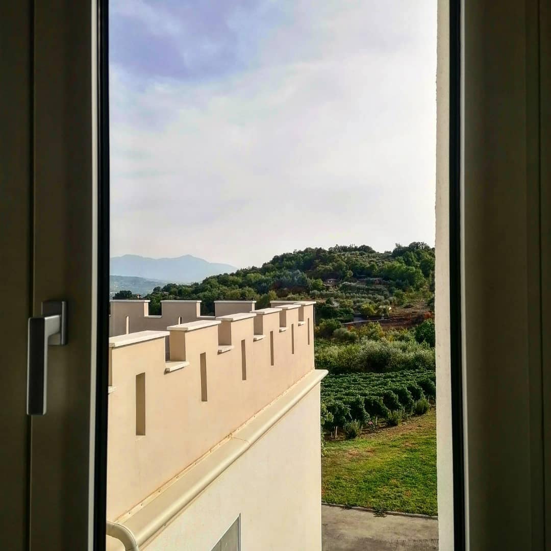 A view from a window showcases rolling green hills and lush vegetation, with a distant mountain visible under a softly clouded sky. The exterior wall features light-colored stucco, harmonizing with the natural surroundings.