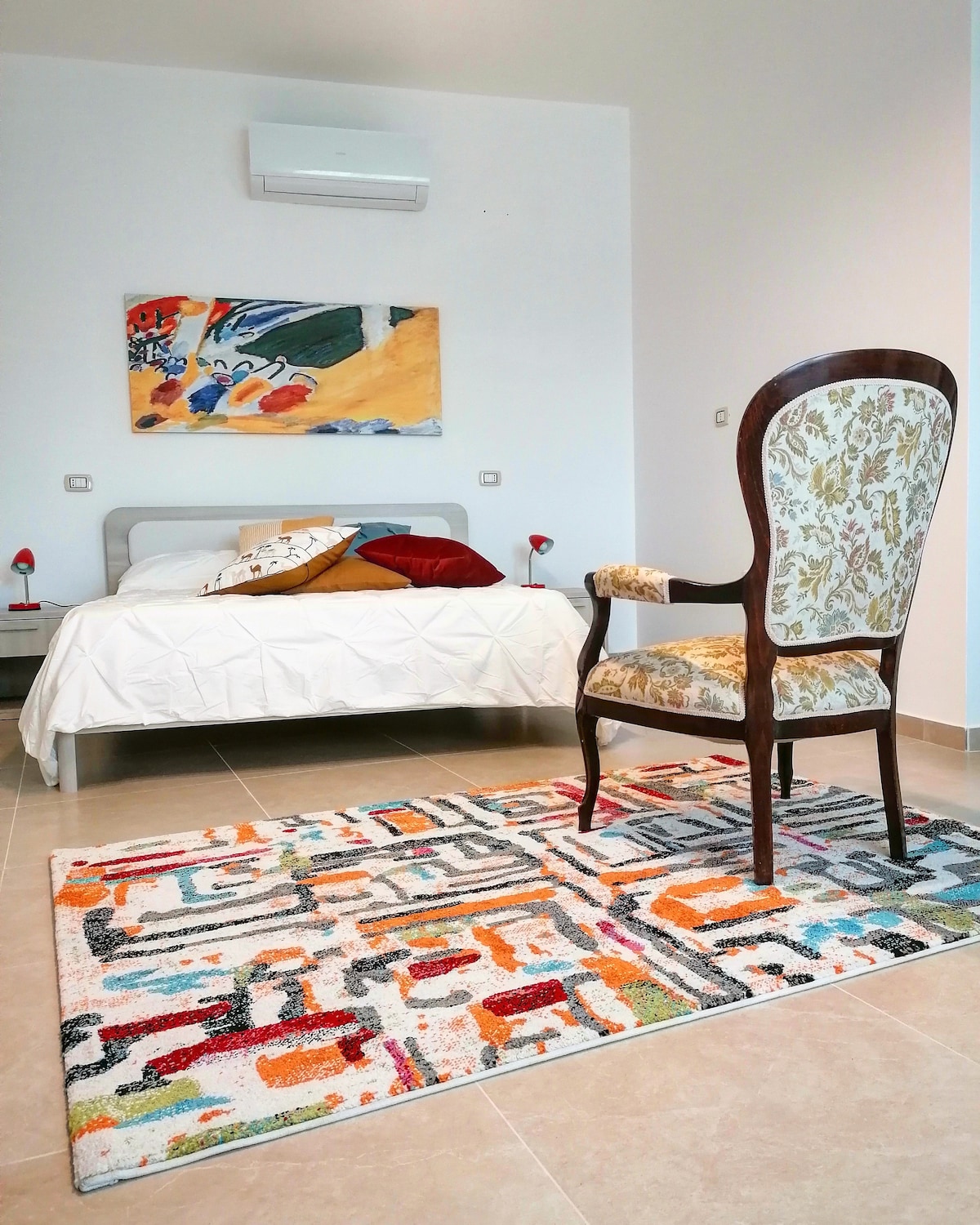 A soothing bedroom features a spacious bed dressed in white linens, complemented by colorful pillows. A decorative chair is positioned nearby, along with a vibrant area rug displaying geometric patterns. The walls are adorned with a contemporary artwork, and air conditioning is installed for comfort.