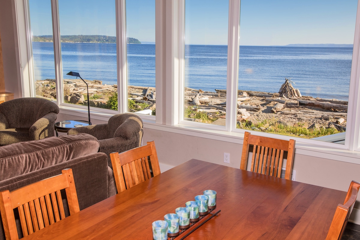 A spacious dining area features a wooden table set for meals, complemented by comfortable seating. Large windows offer expansive views of the calm beach and ocean, allowing natural light to fill the space and showcasing the scenic waterfront.