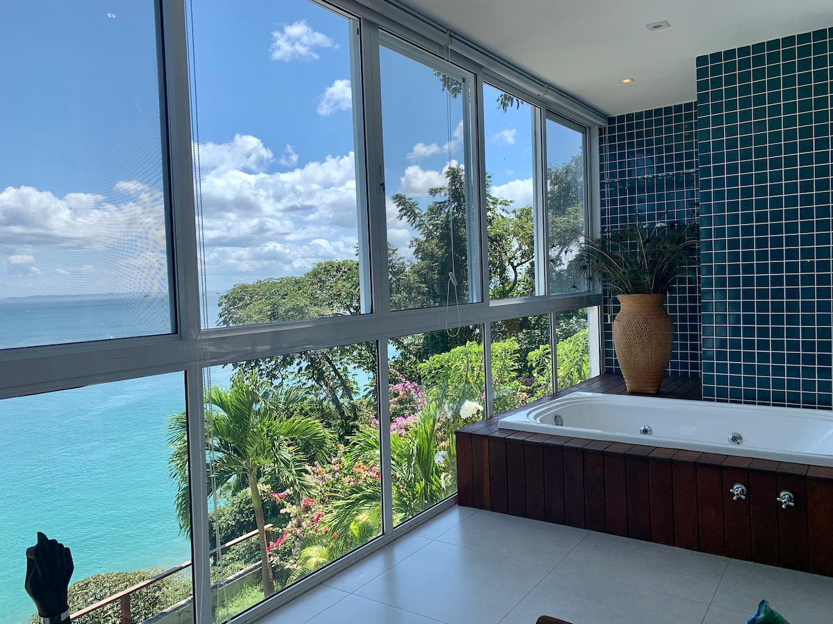 A bright and airy space featuring a jacuzzi positioned next to large windows that showcase a stunning view of the ocean and surrounding greenery. Soft light floods the room, enhancing the calming atmosphere with plants adding a touch of nature.