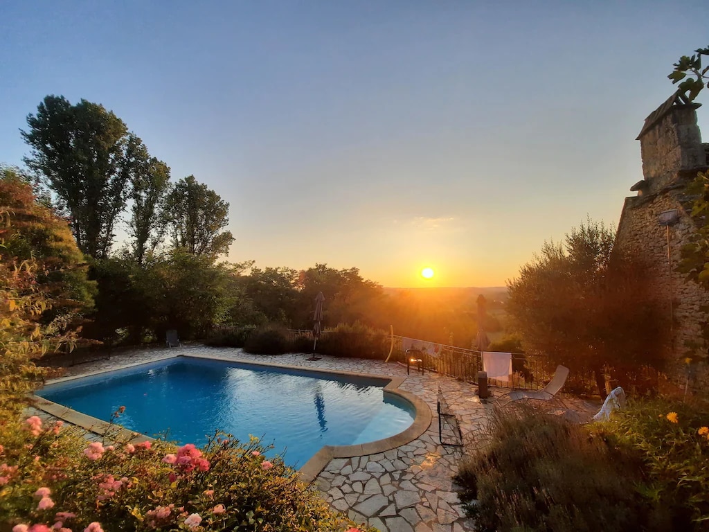 A serene swimming pool is framed by a natural stone patio, with colorful blooming flowers in the foreground. The sky displays warm hues as the sun sets in the distance, casting light over the tranquil setting, while trees provide a lush backdrop.