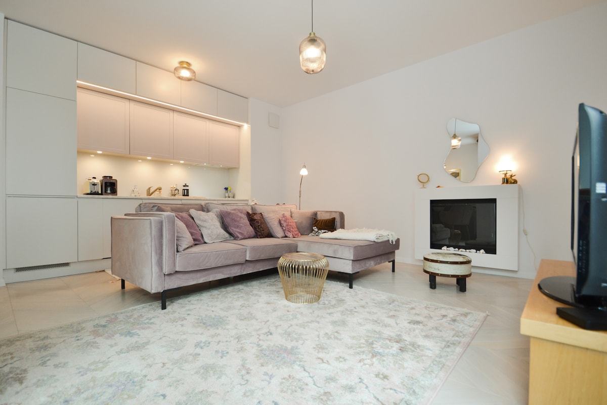 A spacious living area is depicted, featuring a large, soft sofa adorned with decorative pillows. A coffee table sits in front of the sofa, while a television stands on a wooden console. Ambient lighting is provided by modern pendant lamps, creating a calm environment.