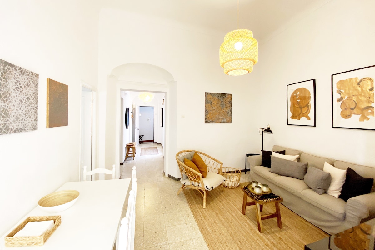 A bright living area is showcased with a light-colored sofa and a woven chair. Artwork in earthy tones adorns the walls. A wooden coffee table holds decorative items, while a neutral rug covers the polished floor, contributing to a spacious and inviting atmosphere.