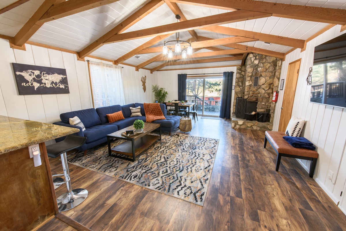 A spacious living area showcases a large blue sectional sofa arranged around a coffee table. A stone fireplace is situated against a wall, while large windows provide natural light. Wooden beams adorn the ceiling, and a patterned rug complements the stylish decor.