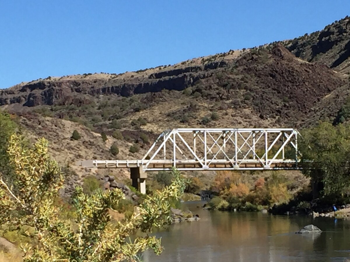 A white metal bridge spans a calm river, surrounded by rocky cliffs and lush vegetation. The landscape features a blend of green and brown hues, with clear blue skies overhead. The tranquil scene reflects a connection to the natural beauty of the area.