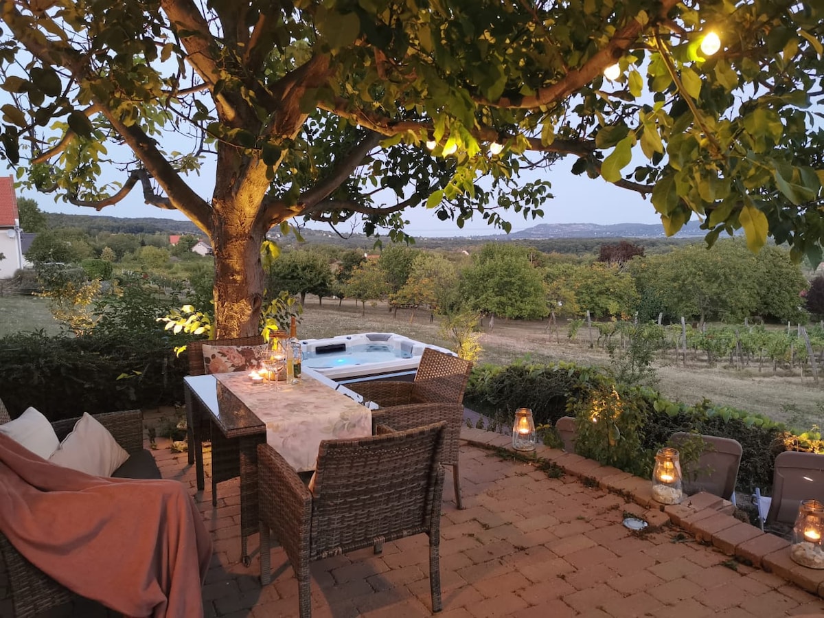 An outdoor dining area is nestled under a large tree, featuring a table and wicker chairs. Soft lighting enhances the ambiance, while a jacuzzi is visible in the background, overlooking a scenic vineyard with rolling hills in the distance.