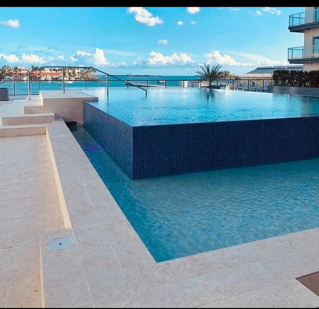 An infinity pool extends toward the horizon, blending seamlessly with the tranquil ocean views. The pool features sleek, modern lines and a tiled edge, complemented by a smooth stone deck. Palm trees are visible in the background against a bright blue sky.