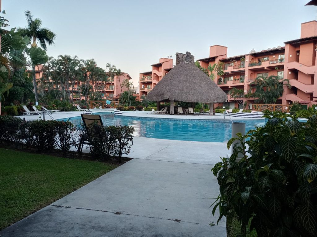 A serene pool area is surrounded by lush greenery, featuring a thatched roof palapa at the water's edge. The pool reflects the surroundings, with various lounge chairs positioned nearby. Colorful resort buildings rise in the background, creating a relaxing atmosphere for guests.
