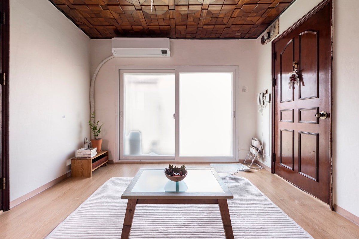 A bright living area featuring a glass table atop a striped rug. A cozy wooden cabinet is positioned beside the sliding door, which leads to an outdoor space. Soft natural light filters in, highlighting the wooden ceiling's texture and creating a spacious feel.