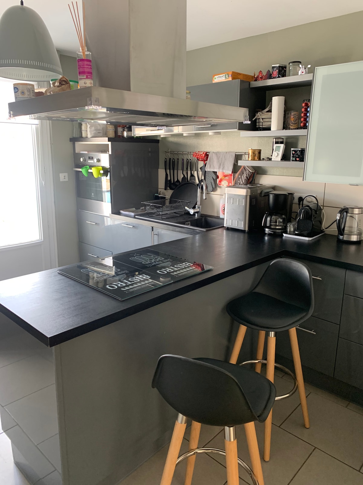 A modern kitchen features sleek cabinetry and a central island with bar stools. Stainless steel appliances, including a coffee maker and kettle, are visible alongside various kitchen utensils. Natural light enters through a window, illuminating the well-organized workspace.