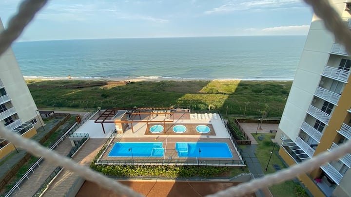 An outdoor area is depicted featuring a swimming pool and several hot tubs, surrounded by green landscape and direct views of the ocean. The horizon meets the water under a clear sky, creating a serene environment just steps from the beach.