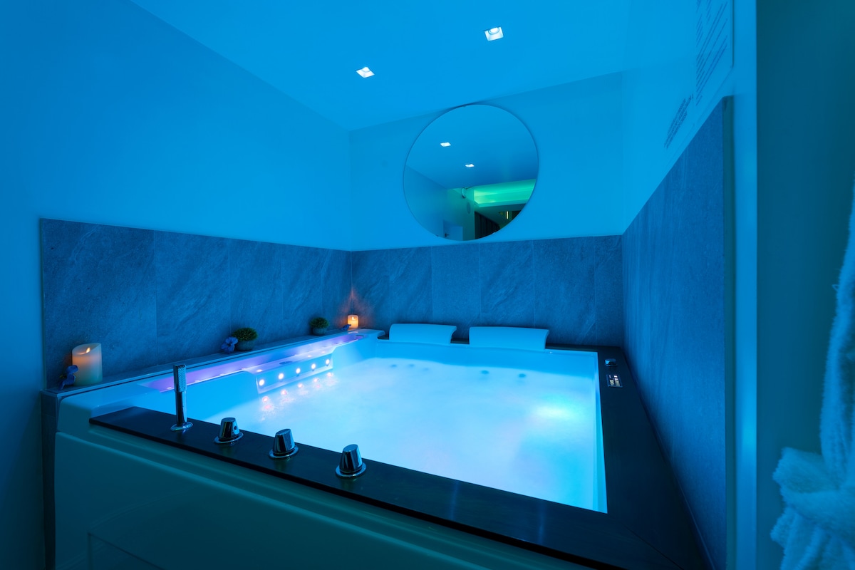 An elegantly designed spa area is illuminated by soft blue lighting, highlighting a spacious hot tub with a sleek edge. Smooth stone tiles cover the walls, while two comfortable loungers sit adjacent to the tub, creating a serene relaxation space.