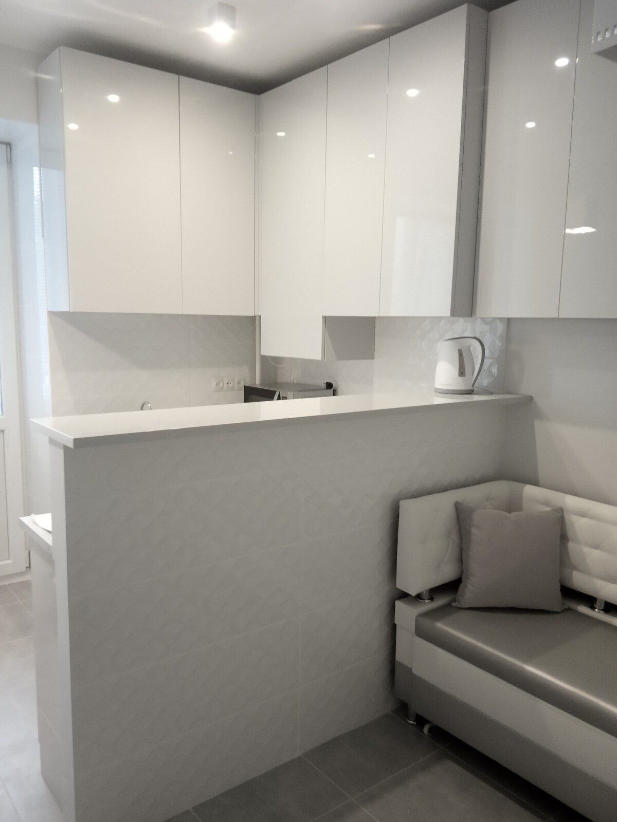 A modern kitchen area features sleek white cabinetry with glossy finishes. A bright countertop is complemented by a small seating nook with a light-colored cushioned bench, accompanied by a gray throw pillow. Natural light is provided by a nearby window, enhancing the clean design.