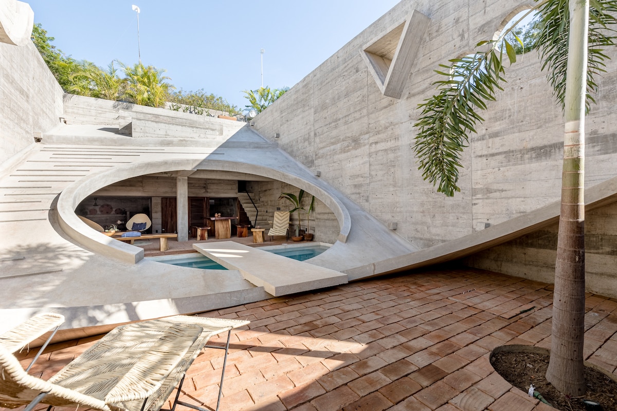 A unique outdoor space is highlighted, featuring a sculptural design with smooth concrete elements. The private pool is integrated within the surroundings, accompanied by a terracotta deck. Lounge chairs provide seating, while palm trees add a touch of greenery to the modern aesthetic.
