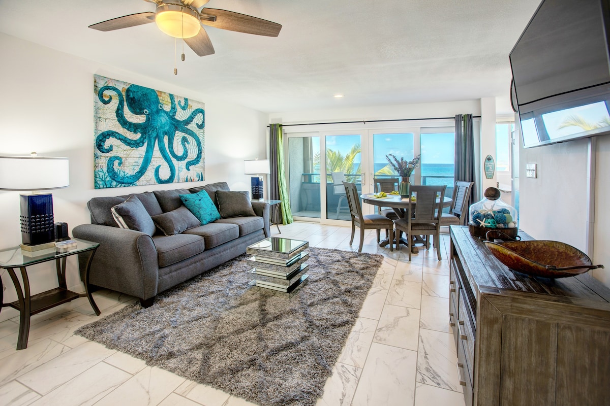 A comfortable living area features a modern sofa and armchair, complemented by a vibrant octopus artwork. Natural light floods through sliding glass doors, revealing ocean views. A dining table set for four and a decorative rug enhance the inviting space.