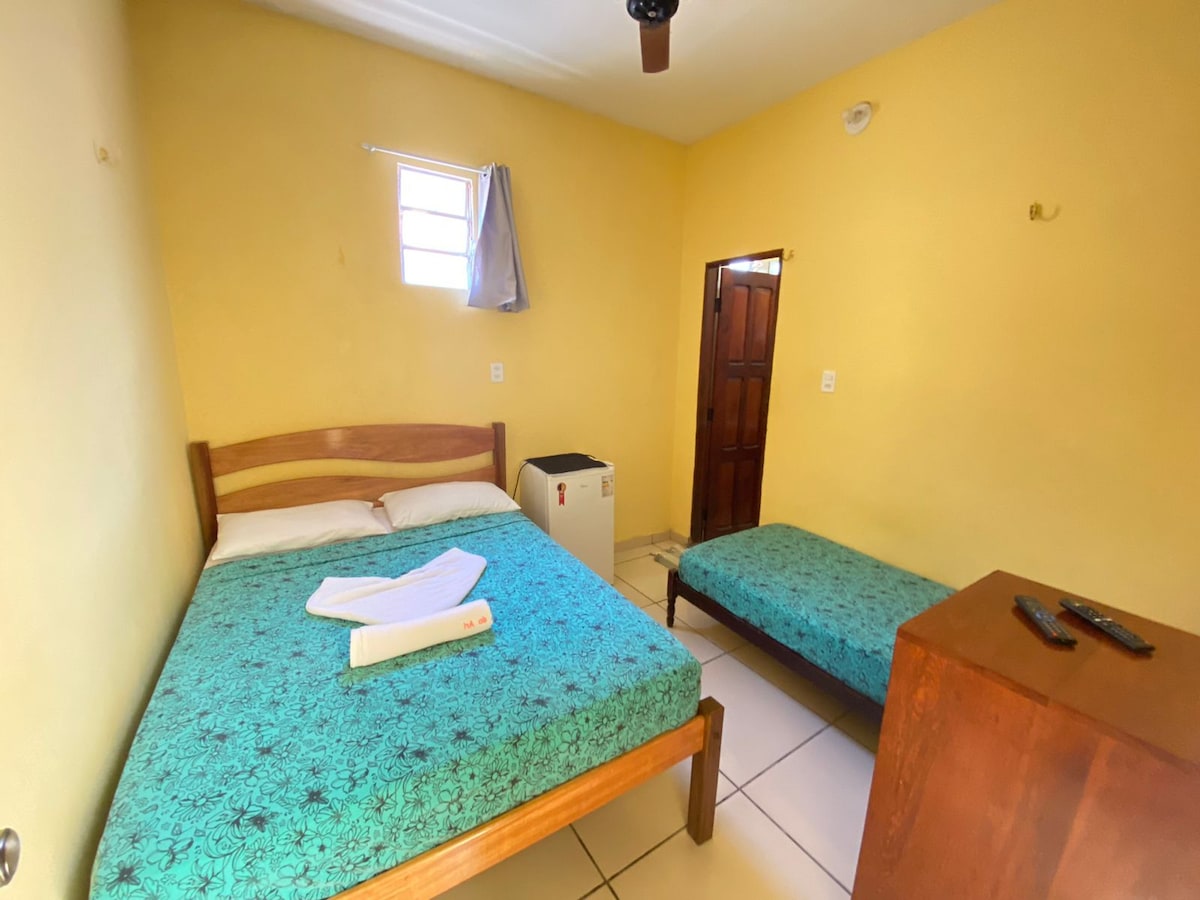 A modest room features two beds, one with a patterned blue cover and the other with a coordinating cover. A small wooden table stands beside the beds, and a door leads to an adjacent space. Natural light enters through a window covered by a light curtain.
