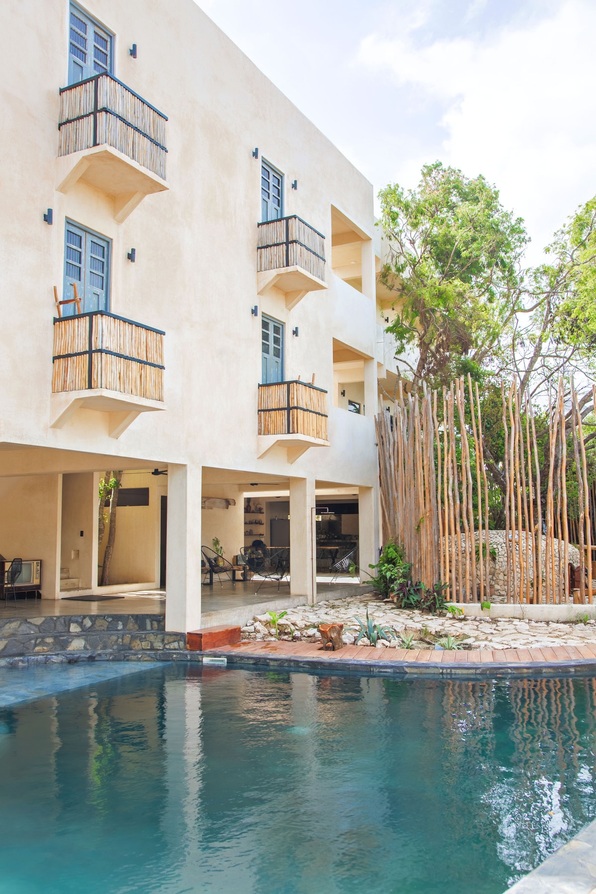 A light-colored building is seen from the outdoor area, showcasing multiple balconies adorned with natural bamboo railings. A serene pool reflects the surrounding greenery, while a decorative arrangement of stones and plants complements the space.