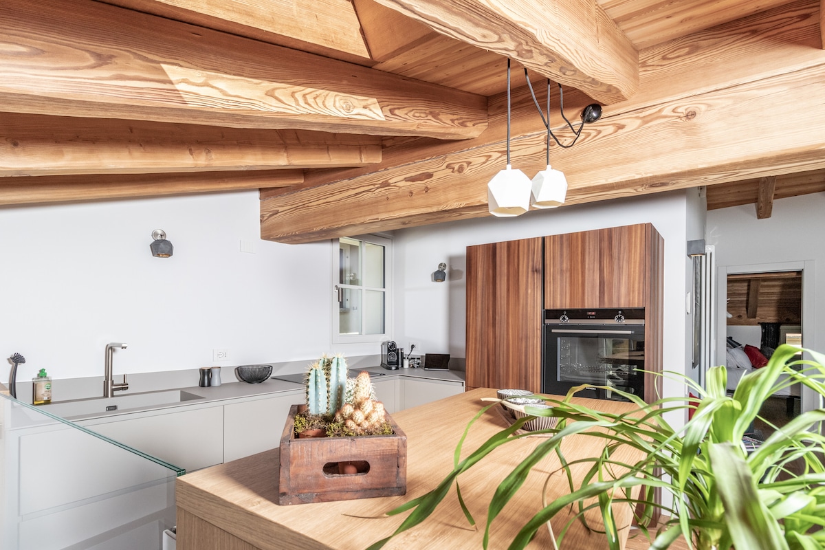 A well-equipped kitchen features wooden beams overhead and modern cabinetry. An island with a wooden surface is positioned centrally, adorned with a decorative planter. A refrigerator and an oven are integrated into the cabinetry, with natural light enhancing the open and inviting atmosphere.