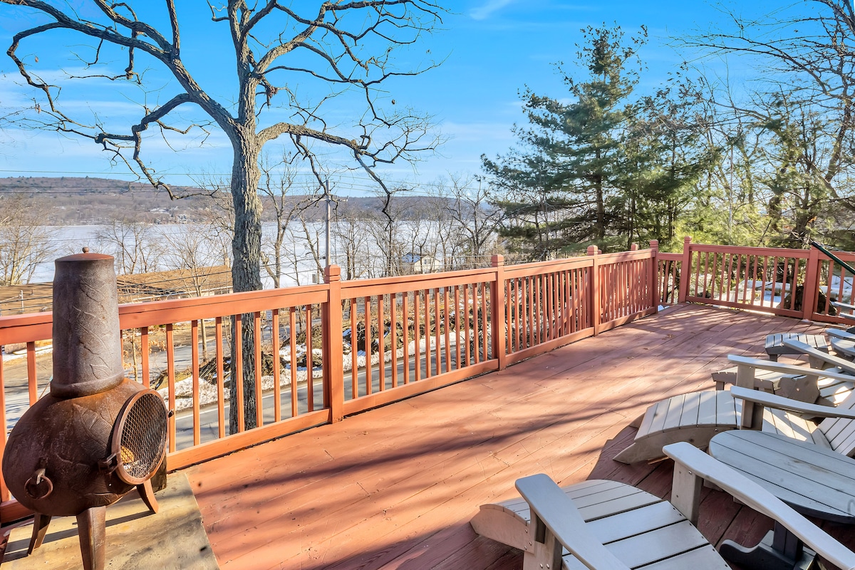 A spacious wrap-around deck is highlighted, featuring multiple white Adirondack chairs facing a scenic view of the lake. A rustic fire pit stands nearby, surrounded by trees that offer a natural backdrop.