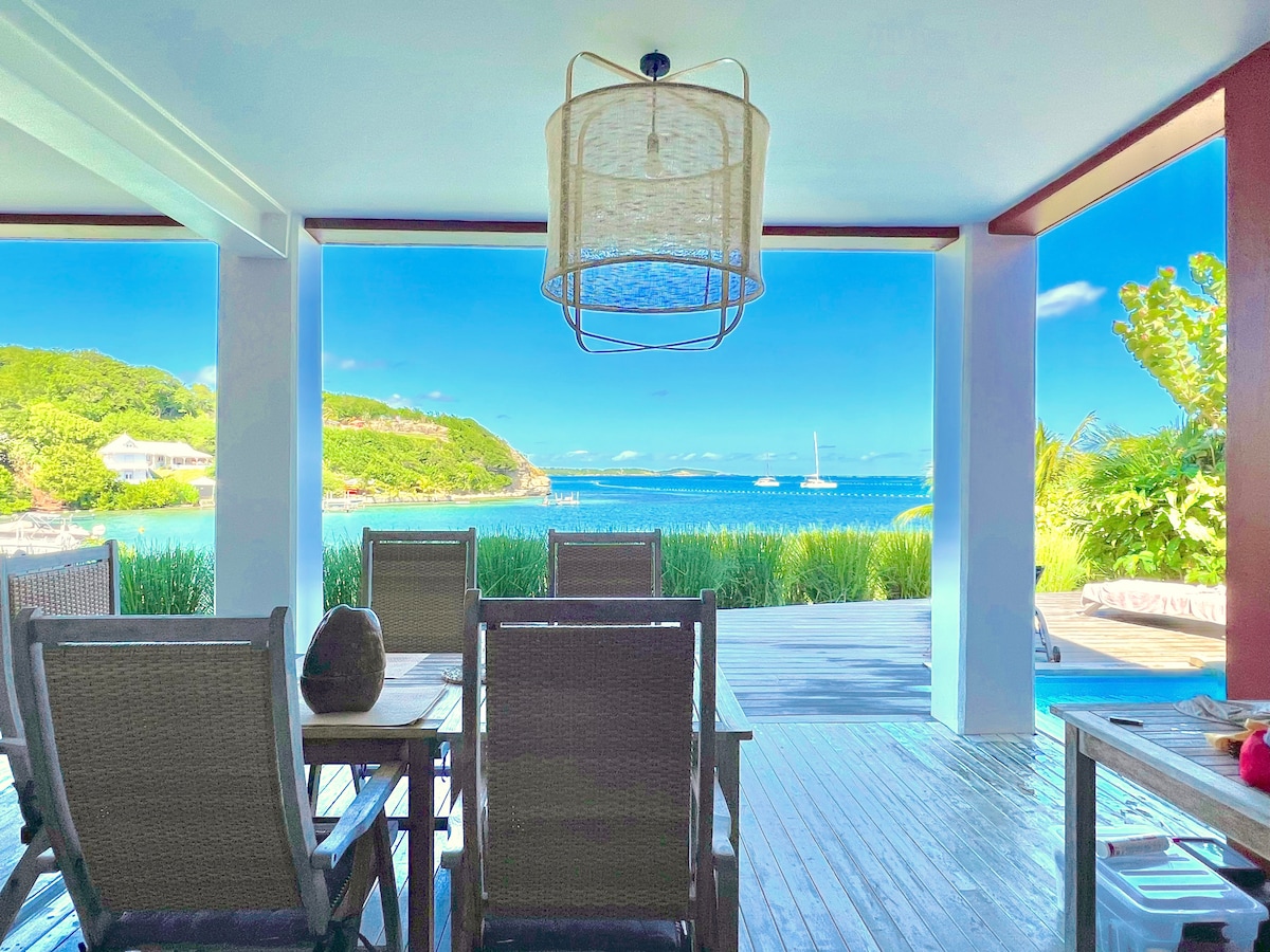 An outdoor dining area features wooden tables and chairs, positioned to face the expansive view of the turquoise lagoon. Lush greenery frames the scene, while a sailboat can be seen on the water. The atmosphere is bright and open, with sunlight illuminating the space.