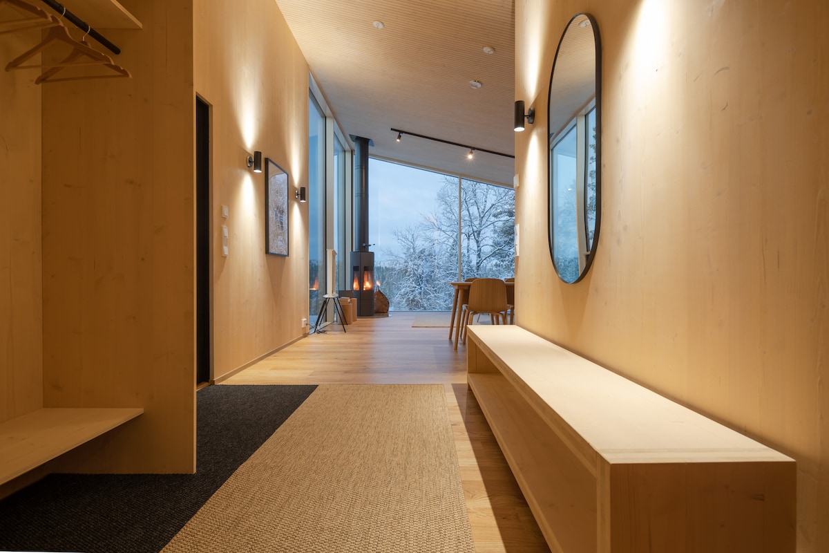 An entryway showcases smooth wooden walls and a spacious wooden bench. A large round mirror reflects natural light. A soft rug runs along the floor, leading to an open area with large windows, providing views of the snowy landscape outside.