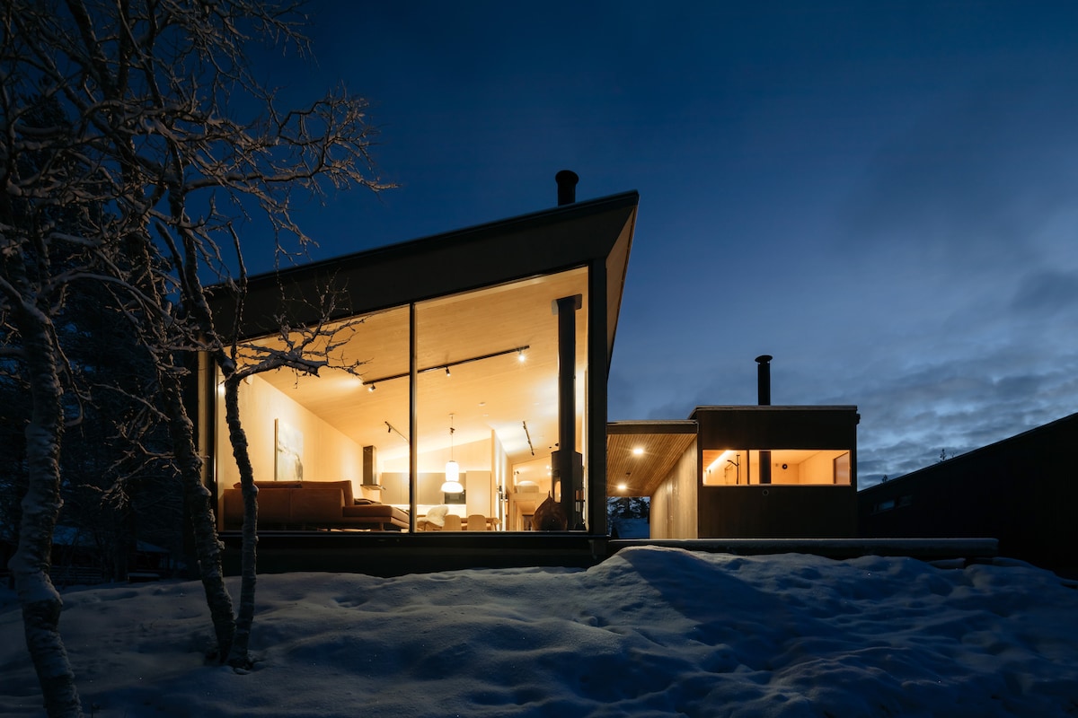 The modern chalet is illuminated at night, showcasing expansive glass walls that offer views of the interior. Snow blankets the ground, and surrounding trees are visible, providing a serene backdrop. The sleek architecture is emphasized by the contrasting warm light from within.