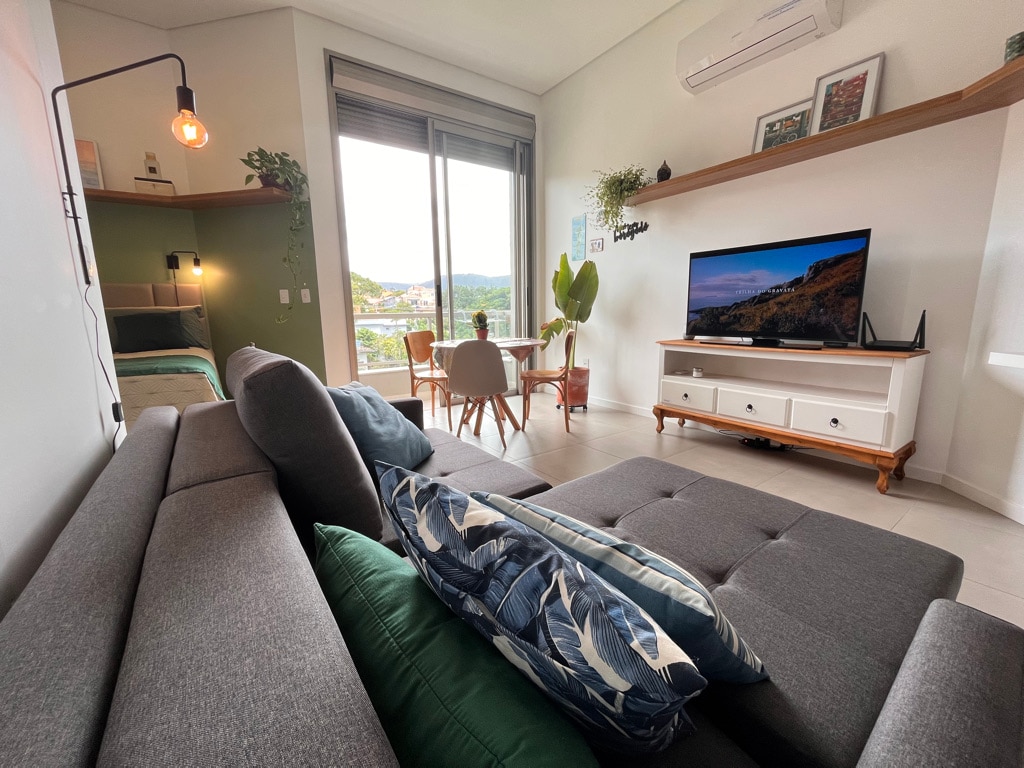 A comfortable seating area features a gray sectional sofa adorned with leaf-patterned cushions. A small dining space with light wooden chairs is visible, and a flat-screen TV is mounted on a white cabinet. Large windows provide ample natural light and scenic views of the surroundings.