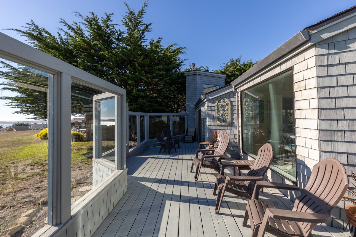 An enclosed ocean-facing deck is shown, featuring several wooden lounge chairs arranged for relaxation. Large glass windows provide unobstructed views of the surrounding landscape, with trees visible in the background under clear skies.