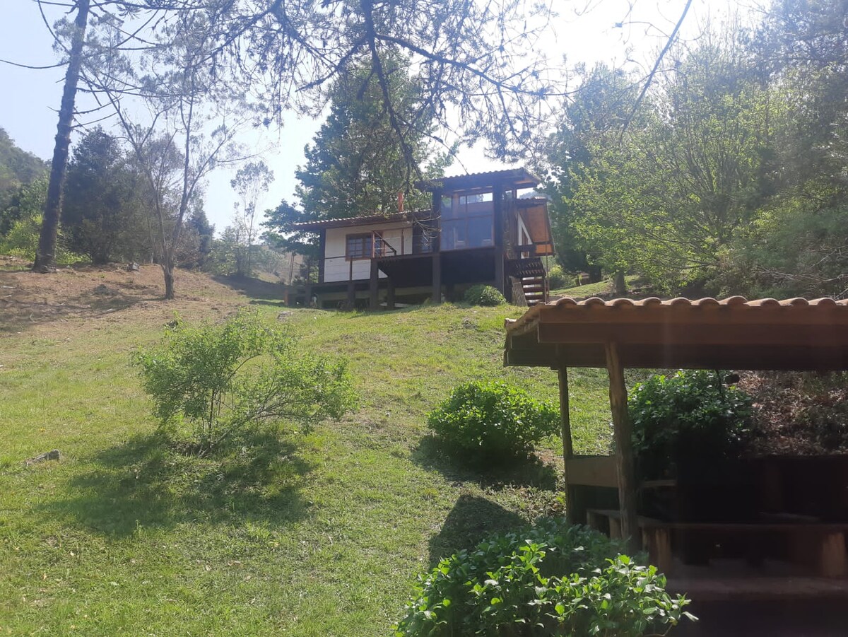 A cozy chalé is set amidst a lush green landscape, surrounded by trees. A wooden gazebo with a roof is visible in the foreground, while the chalé features large windows and a deck. Natural sunlight illuminates the scene, highlighting the peaceful environment.