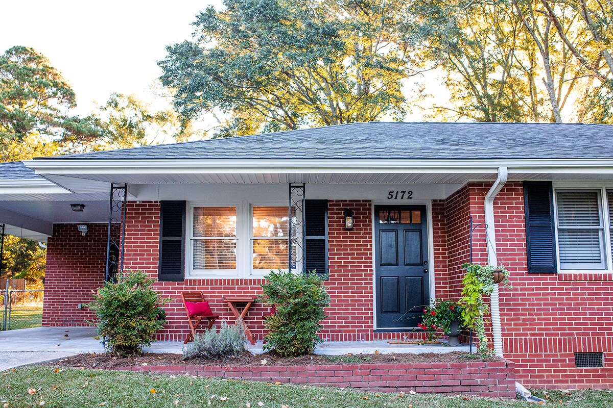 The vintage brick facade of the house is complemented by well-maintained landscaping. A small wooden table with two red chairs is positioned by the front door, framed by greenery. Sunlight filters through the trees, casting soft shadows on the porch.