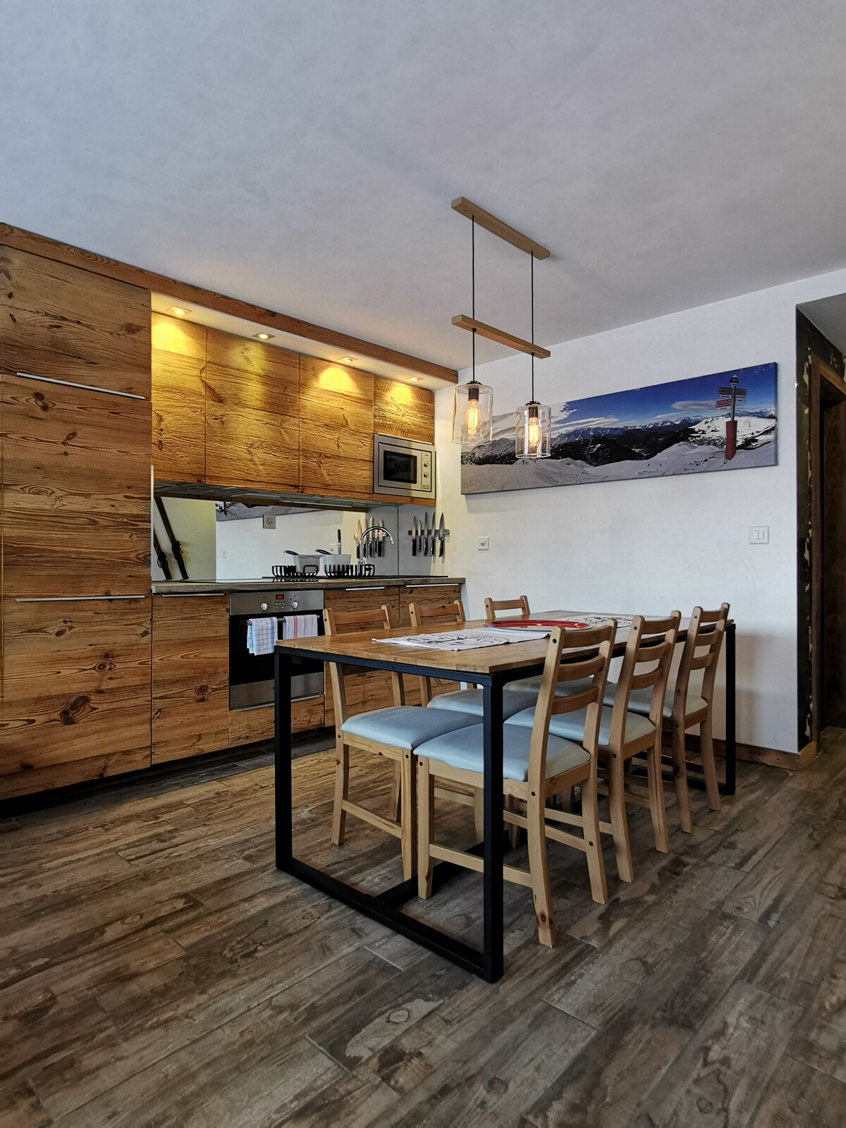 A modern kitchen area features wooden cabinets paired with sleek appliances, including a microwave. A dining table with minimalist chairs is set for meals, while artwork depicting mountain scenery is displayed on the wall. Warm lighting enhances the inviting ambiance.