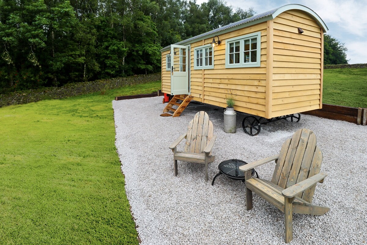 A quaint shepherd's hut is set on a gravel patio, featuring two rustic wooden chairs arranged around a small fire pit. Lush greenery surrounds the hut, and a set of steps leads to a bright entrance, showcasing multiple windows that provide natural light.
