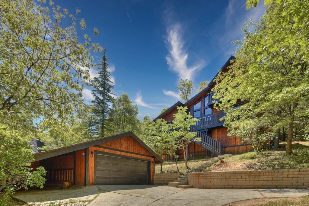 The exterior of a chalet is shown, complete with a two-car garage and a sloped driveway. Surrounded by lush green trees, the wood-paneled façade is complemented by a blue balcony overlooking the natural landscape. A clear sky enhances the inviting atmosphere.