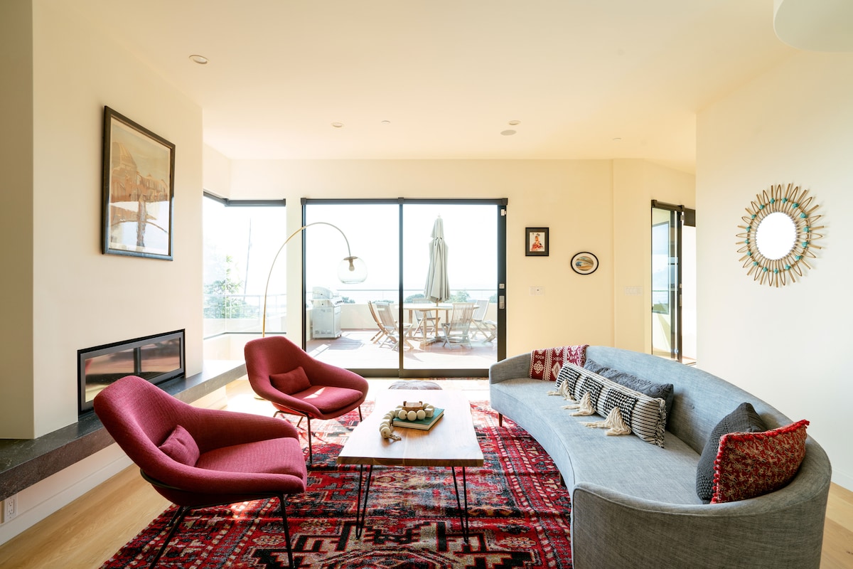 An open living area is featured, showcasing a curved gray sofa and two vibrant red chairs. A low coffee table sits in the center, adorned with decorative items. Large glass doors lead to an outdoor deck, allowing natural light to fill the space.