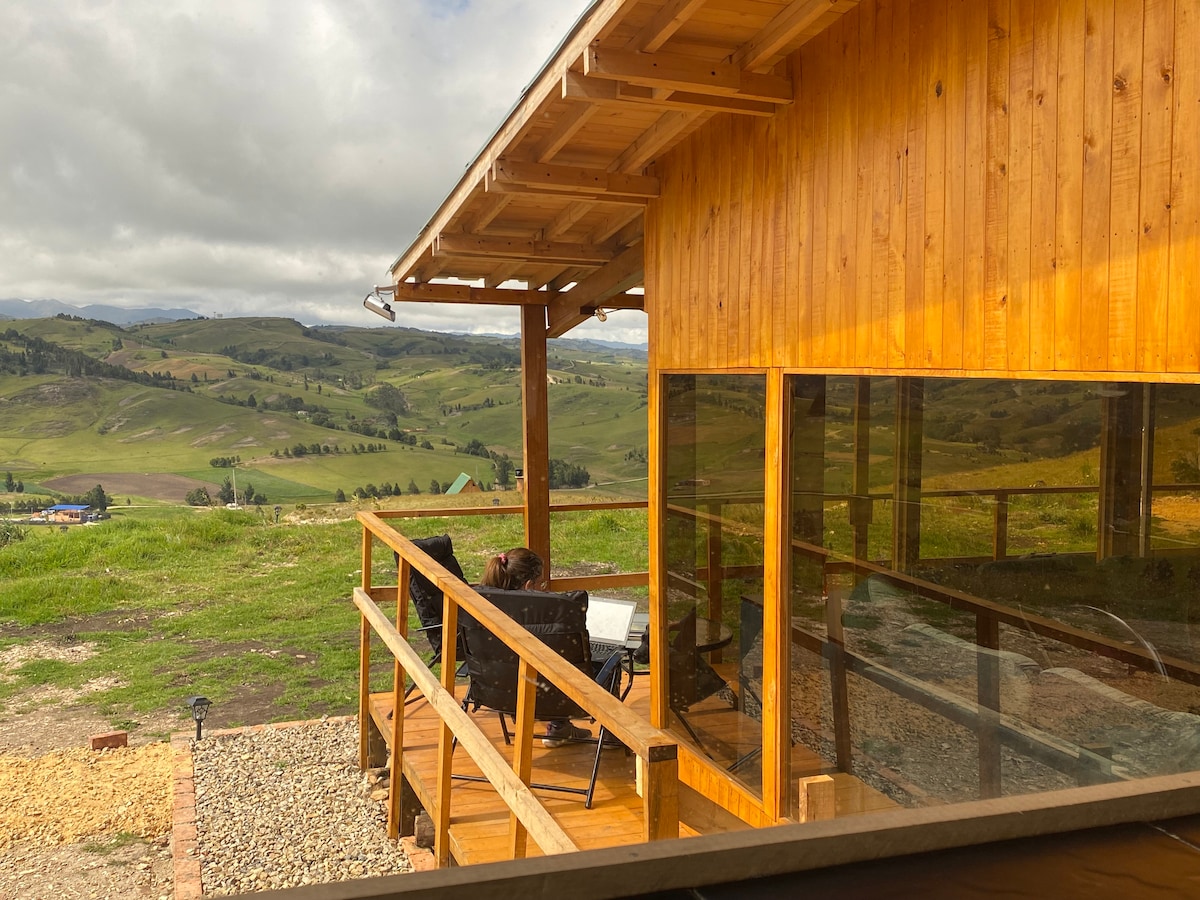 A wooden deck offers seating with a clear view of the surrounding hills. Large glass windows provide natural light and showcase the tranquil landscape, while the rustic architecture complements the natural setting.