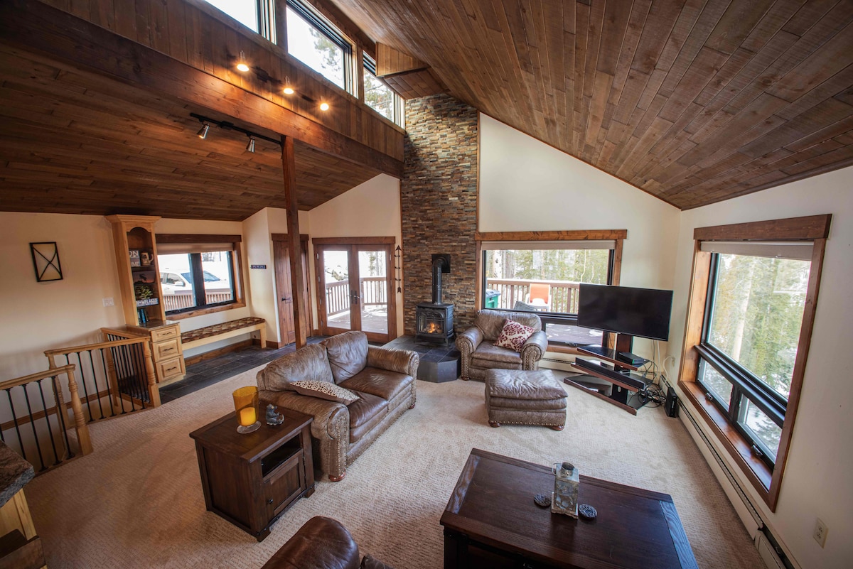 The main living area is characterized by vaulted ceilings and an open floor plan. Floor-to-ceiling windows provide natural light and panoramic views. A gas fireplace and a large television are positioned for comfort, complemented by leather seating and warm wooden accents throughout.