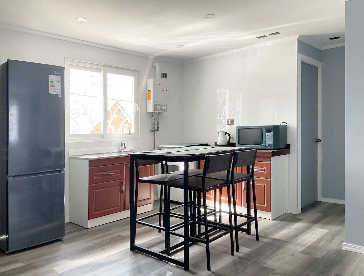 An equipped kitchen area features a tall dining table with black chairs, a refrigerator, microwave, and a small stovetop. Natural light fills the space through a window, illuminating the clean countertops and modern cabinetry in warm hues.