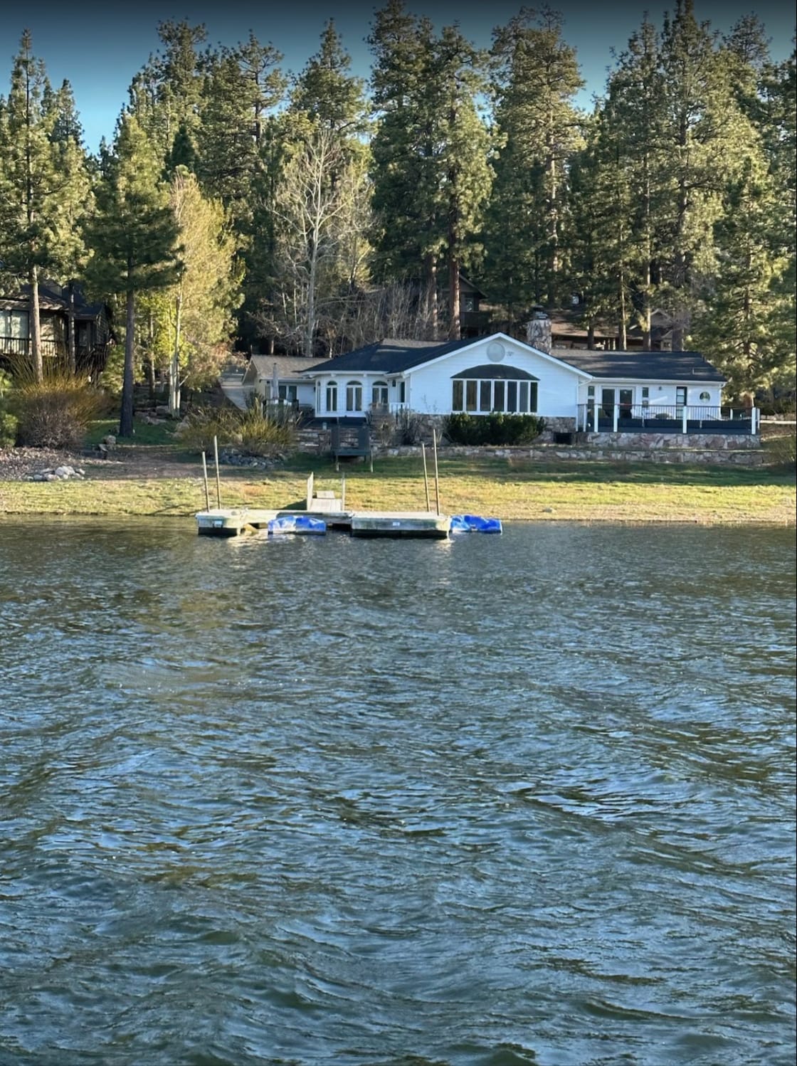 The exterior of the cabin is visible from the water, showcasing large windows that reflect the surrounding trees. Two floating docks are anchored nearby, providing a perfect spot for swimming and boating. The scenery is complemented by tranquil lake waters and lush greenery in the background.
