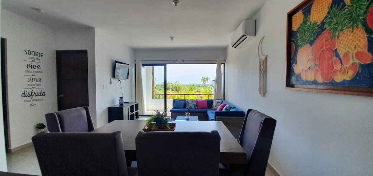 A modern dining area is presented with a rectangular table surrounded by four upholstered chairs. The space opens to a living area featuring a comfortable couch and a large window offering a view of the outdoors. A colorful wall art piece enhances the decor.