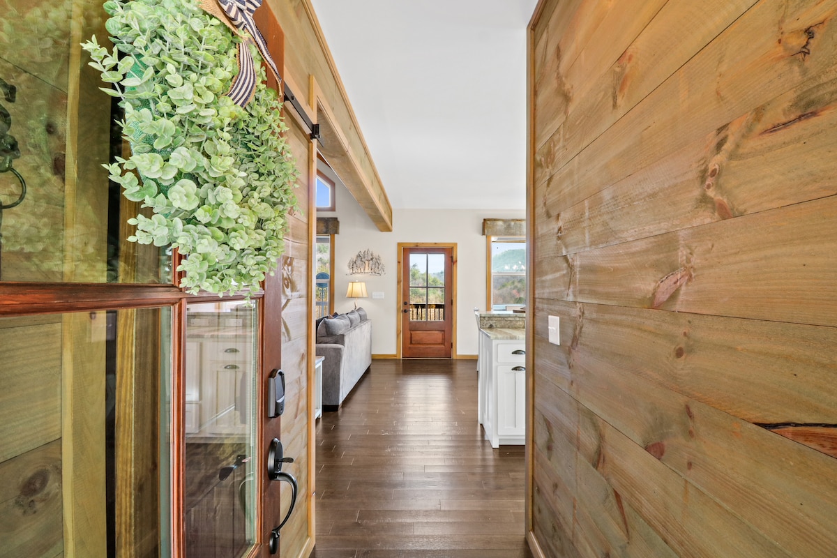 A welcoming entrance showcases rustic wood walls and a bright green hanging plant. The space leads into an open area with natural light streaming through the glass front door, revealing glimpses of seating and kitchen access through the archway.