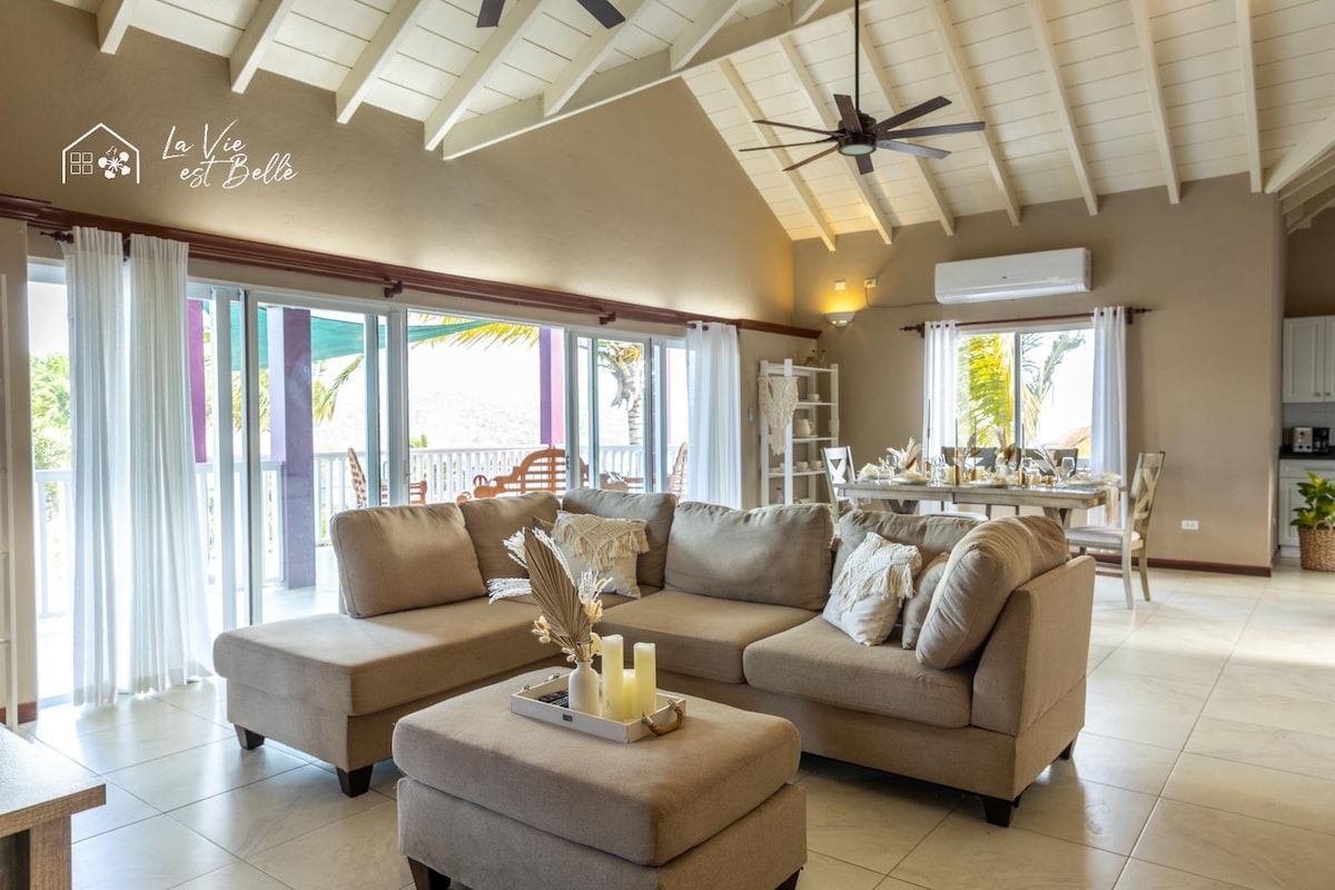 A spacious living area features a large, neutral-toned sectional sofa with decorative cushions. Bright, retractable glass doors provide access to a terrace, enhancing natural lighting. A dining table is set in the background, complemented by light wood accents and a ceiling fan.