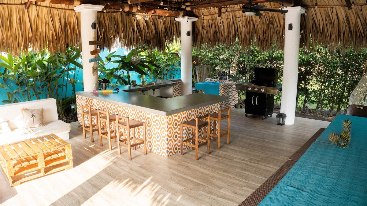 An outdoor rancho area is highlighted, featuring a spacious bar counter with patterned tiles and several wooden stools. The setting includes a barbecue grill and comfortable lounge seating. Lush greenery surrounds the space, with natural light enhancing the inviting atmosphere.