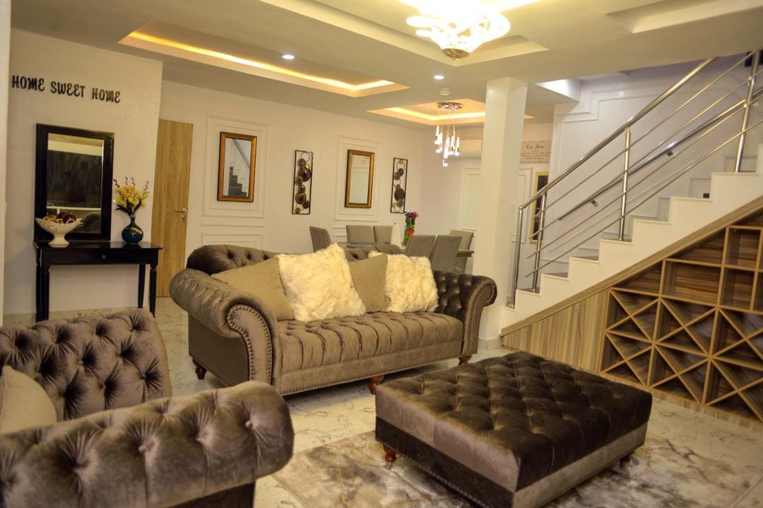 A cozy living area is furnished with plush sofas and stylish ottomans, complemented by a central coffee table. Elegant wall art and decorative accents create a welcoming ambiance, while soft lighting enhances the room's openness. A staircase leads to the upper level, adding architectural interest.