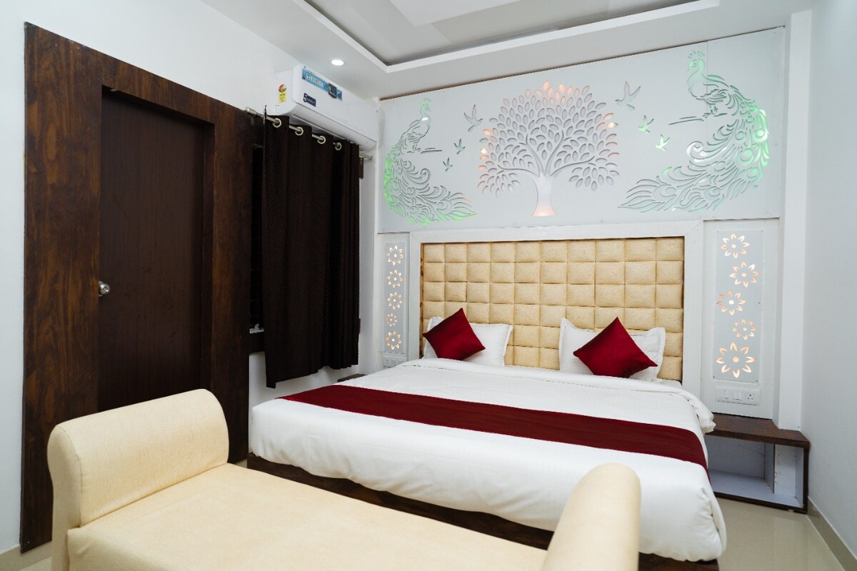 The room features a spacious bed with a cushioned headboard and decorative red accents. A subtle wall design of peacocks adds a unique touch. A light-colored seating area is positioned nearby, complementing the overall decor. Natural light filters through a window covered by dark curtains.