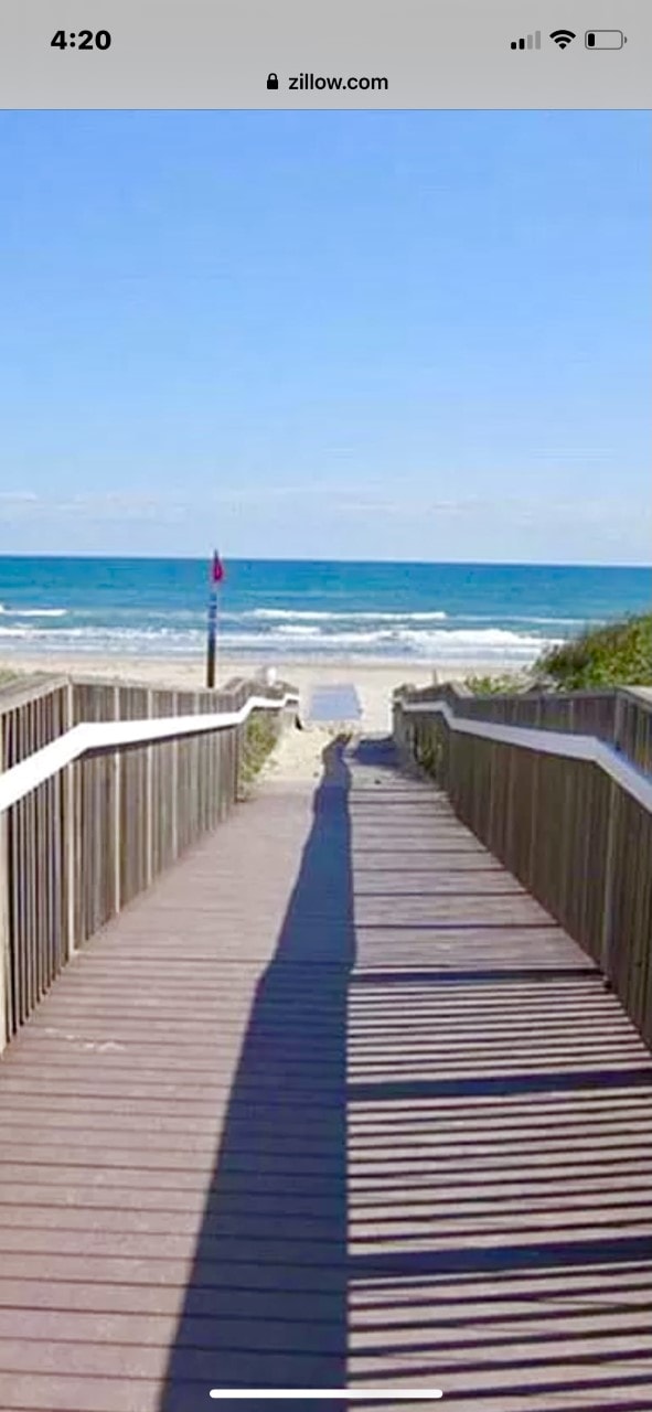 A wooden boardwalk leads directly to the sandy beach, framed by greenery on both sides. The ocean is visible at the end of the path, featuring calm turquoise waters and a bright blue sky scattered with few clouds.